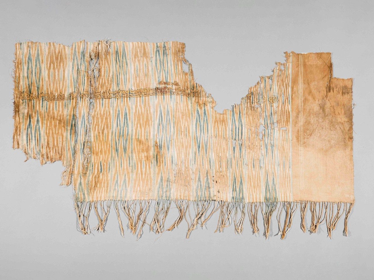 Blue and tan patterned textile with gold a line of text across the upper portion and fringe along the bottom.