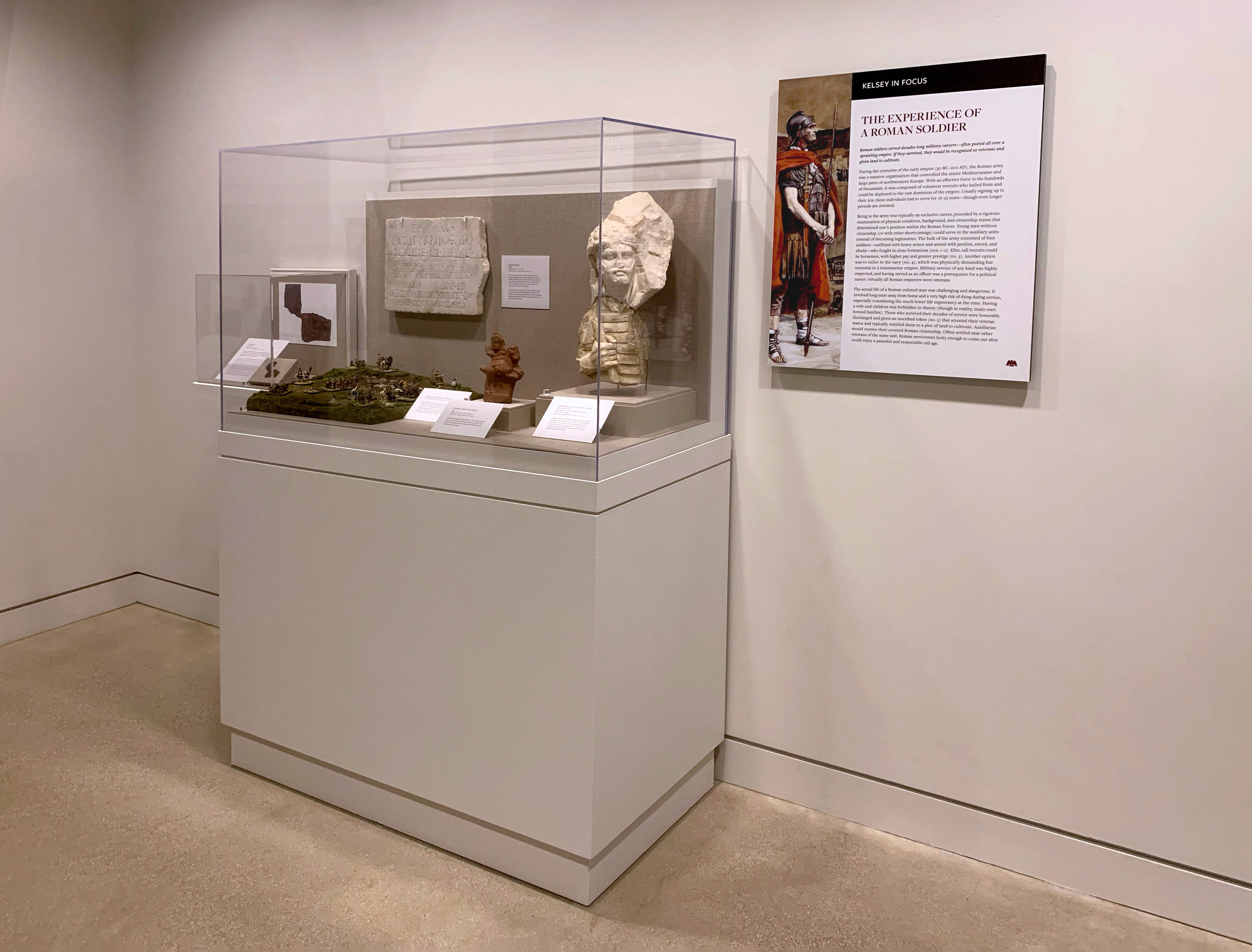 Museum vitrine showing a diorama of soldiers on a battlefield, a marble tombstone, a clay horseman figurine, and the head and soldier of a Roman soldier. On the wall next to the case is a panel titled “The Experience of a Roman Soldier.”