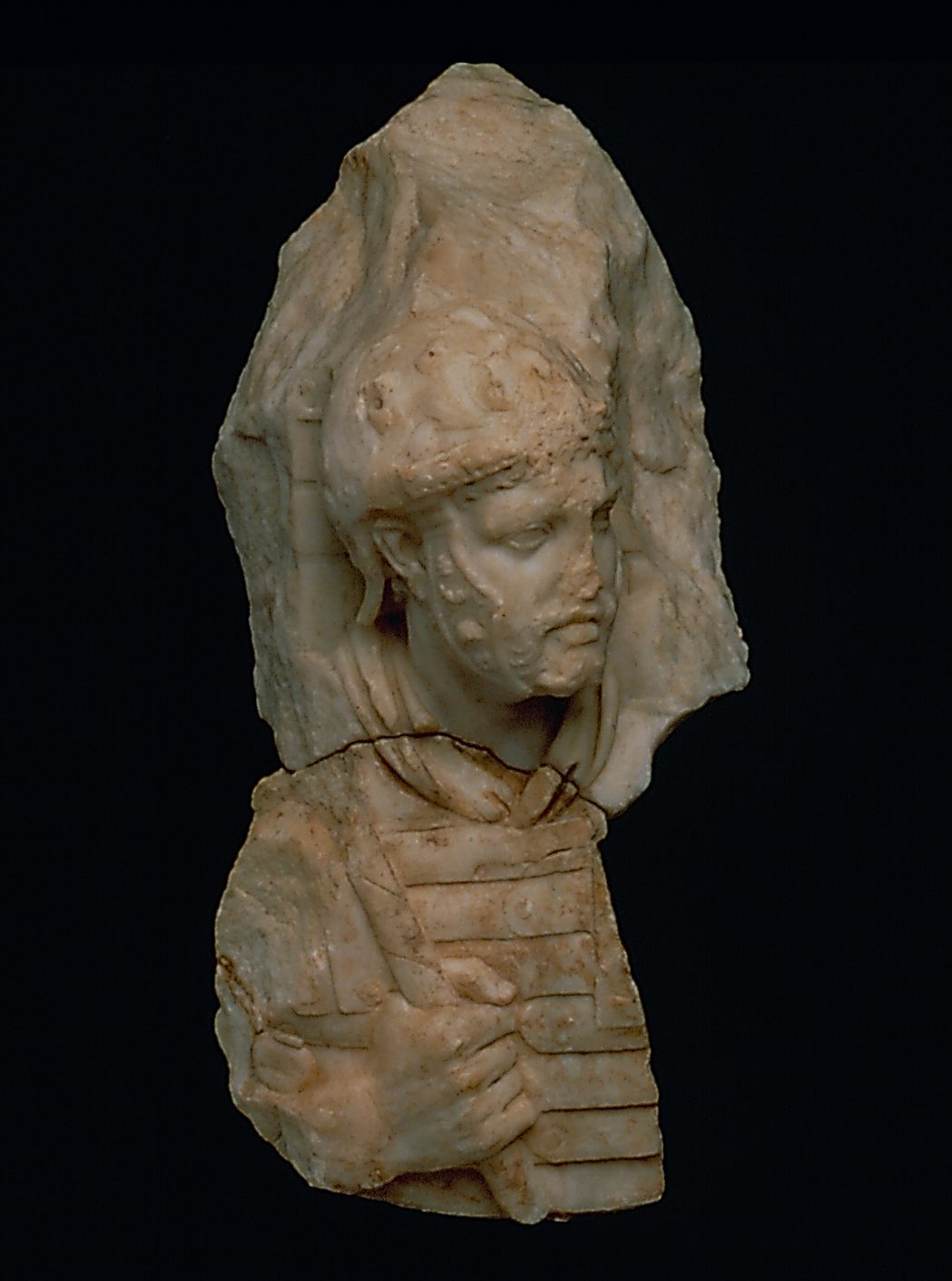 White marble relief figure of a helmeted head of a male soldier and a torso with a hand holding part of a spear. The head and torso are broken apart from one another. 