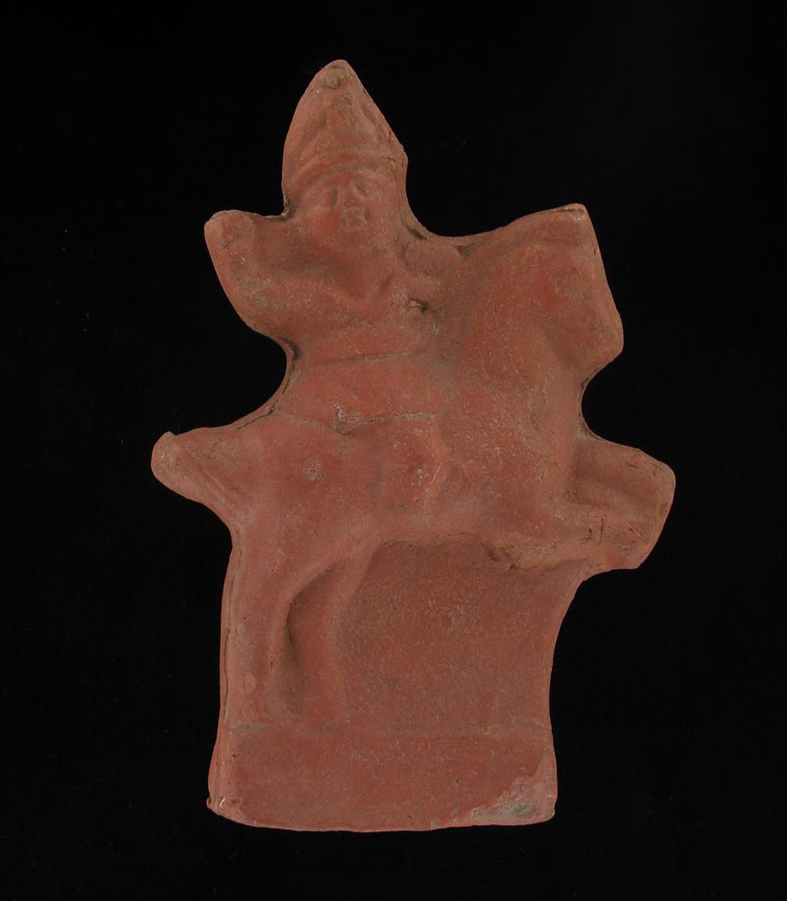 A red clay artifact against a black background. The figurine wears a helmet, lifts a hand aloft, and rides a horse with raised forelegs.