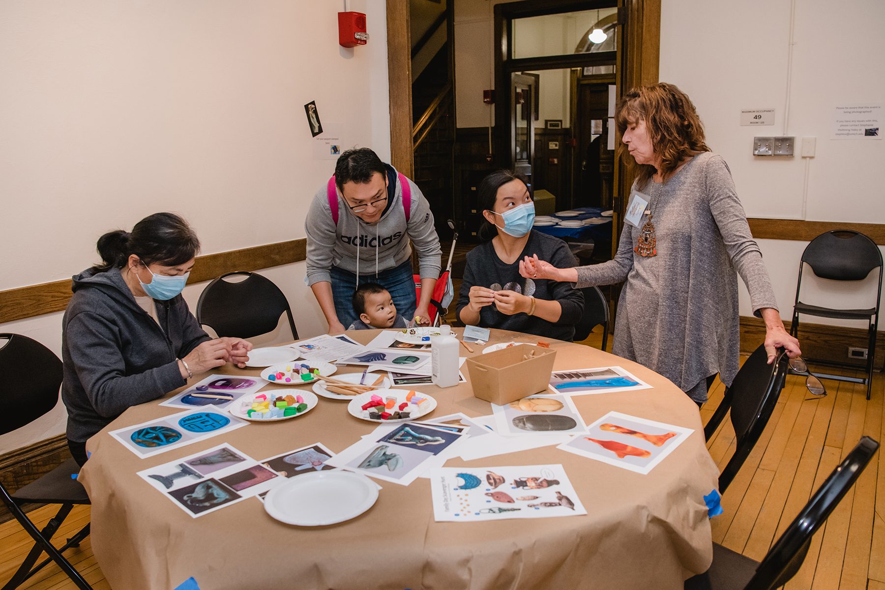 Three adults and a toddler at a table covered with a paper tablecloth, reference photos of Kelsey Museum artifacts, and clay to make crafts. A volunteer docent speaks with one of the adults at the craft station.