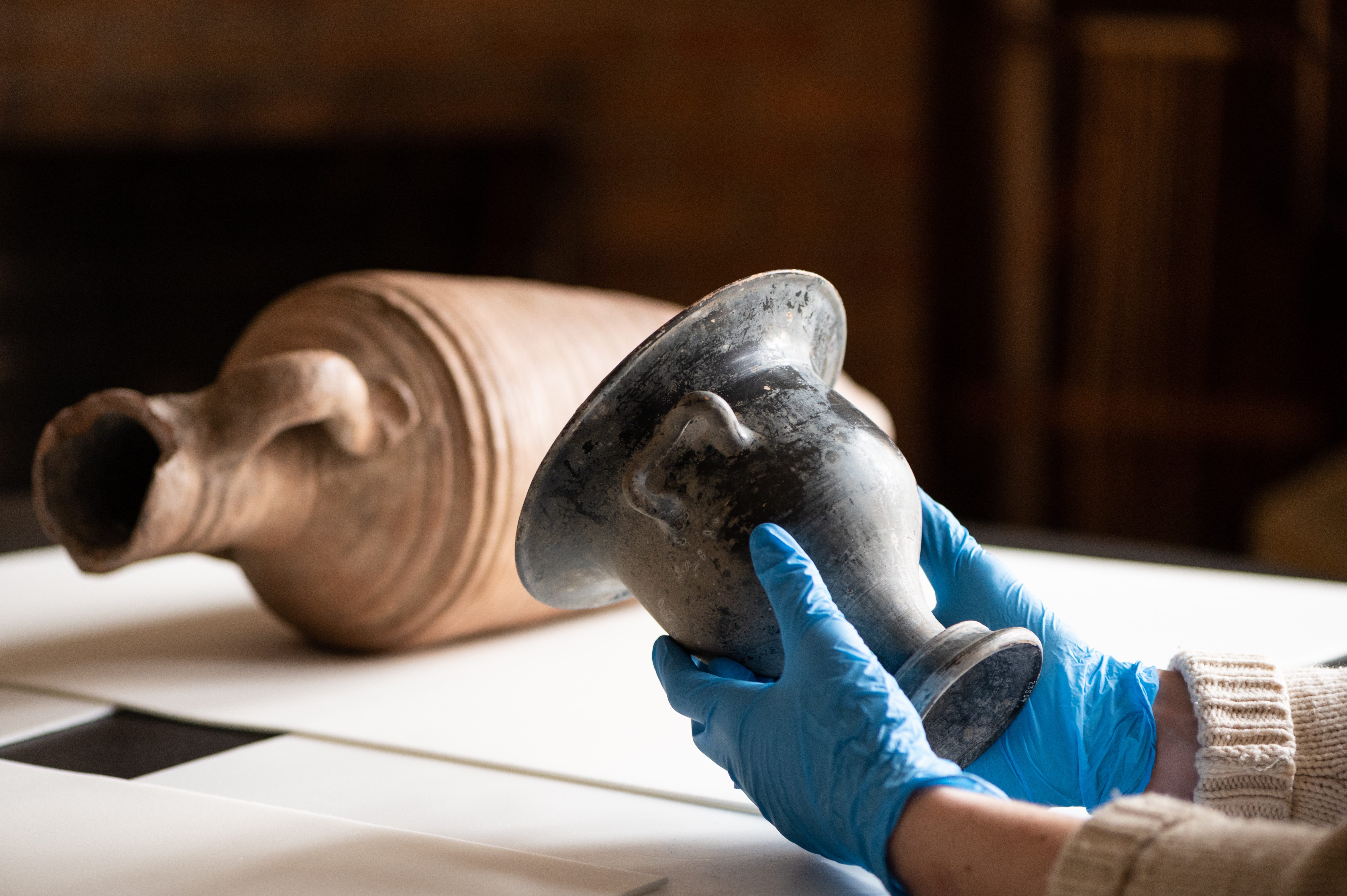 A person wearing blue gloves holds a black vessel. In the background, a buff amphora lies on its side. 