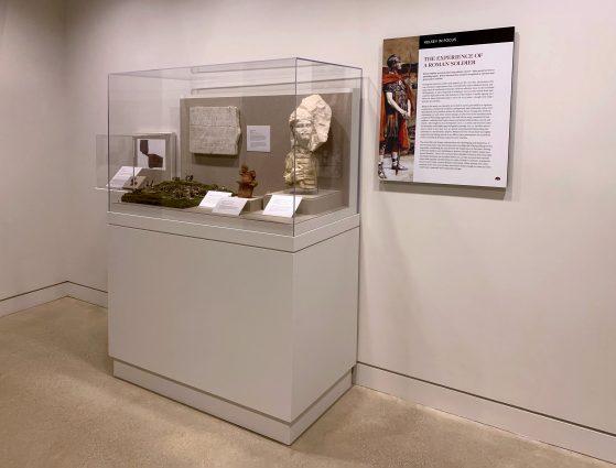 Museum vitrine showing a diorama of soldiers on a battlefield, a marble tombstone, a clay horseman figurine, and the head and torso of a Roman soldier. On the wall next to the case is a panel titled “Kelsey in Focus: The Experience of a Roman Soldier.”