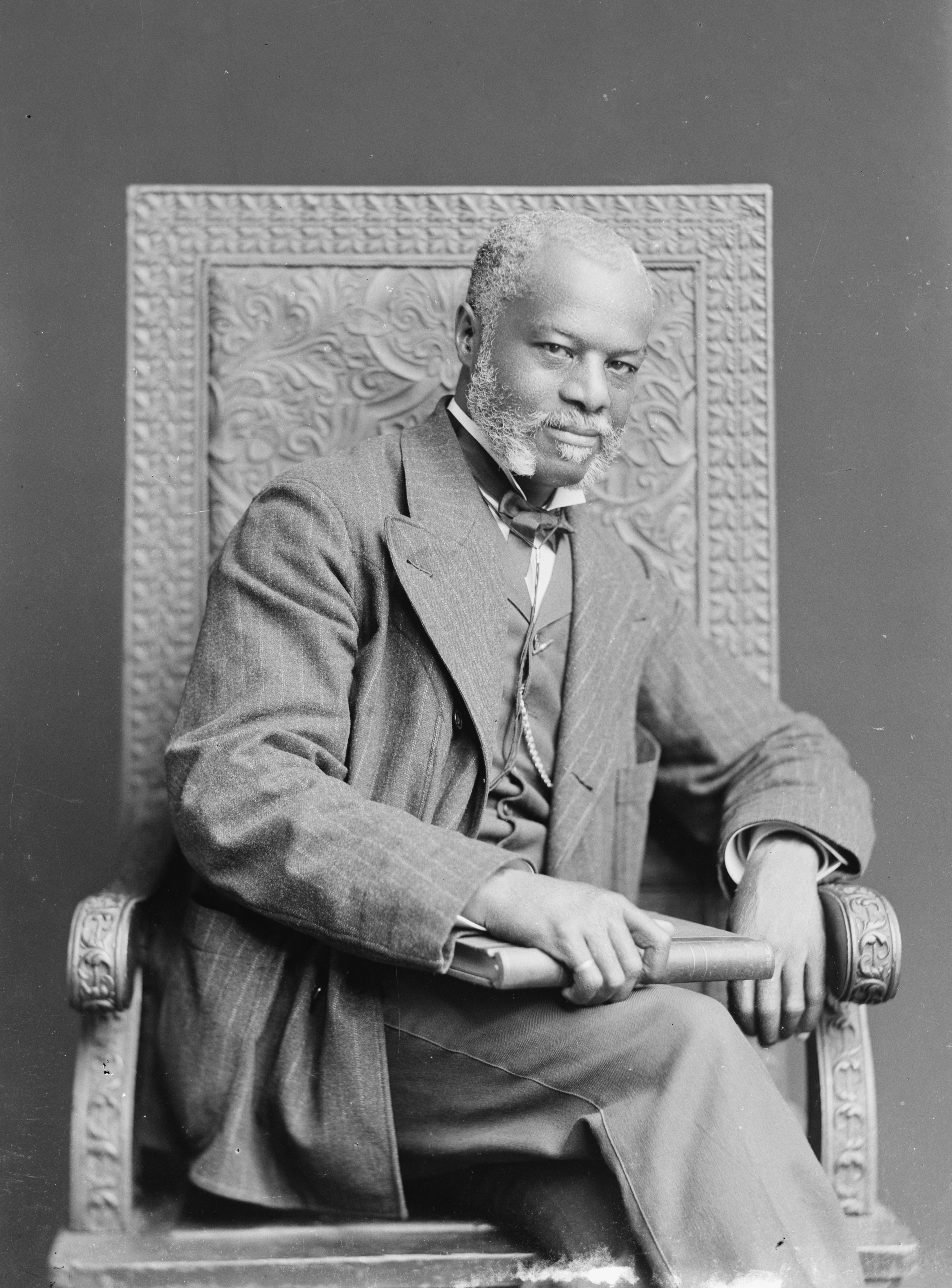 Bearded African American man holding a book, sitting in a decorative chair for a portrait. 