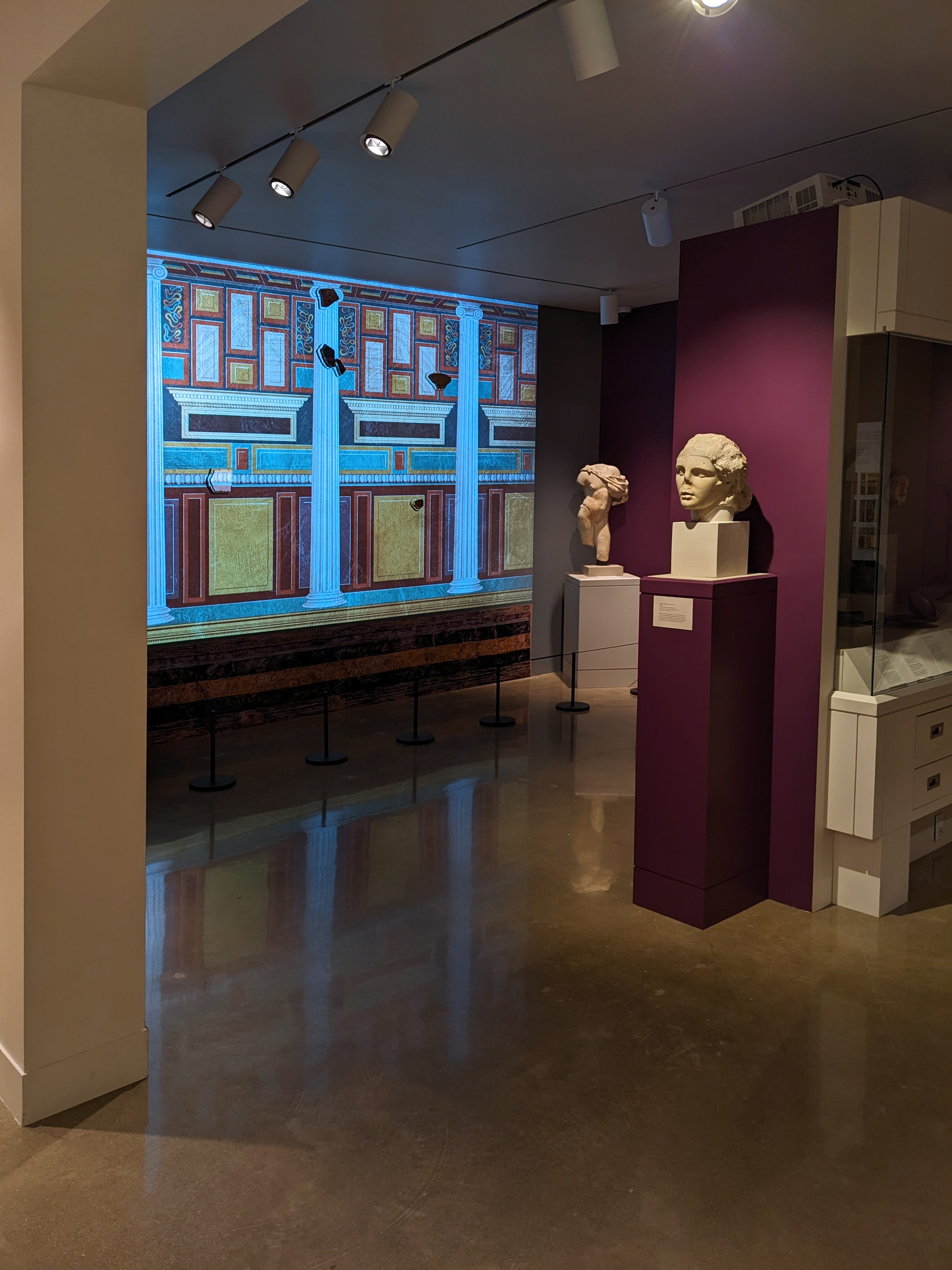 View into the Kelsey Museum’s Roman galleries. A large image of a Roman wall painting, featuring columns and other architectural elements, is projected onto the rear wall. White marble statues on pedestals are also visible in the gallery space. 