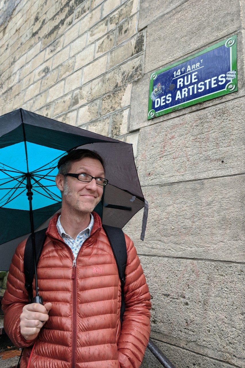 Bruce Worden, holding an umbrella, looks up at a sign reading, “Rue Des Artistes.”