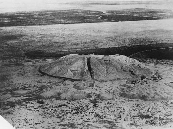 Black and white aerial view of a large earthen mound in a desert context. 