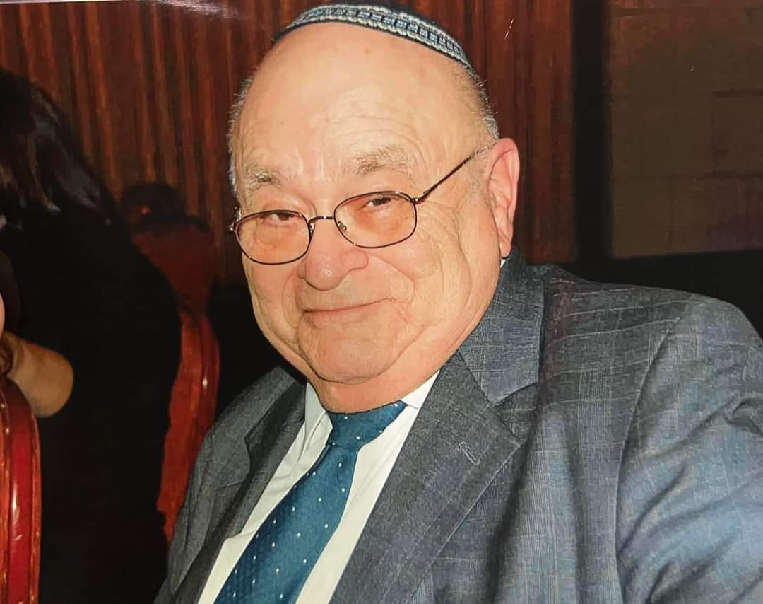 A smiling candid picture of Gene Moshe Schramm wearing a grey suit and teal tie.