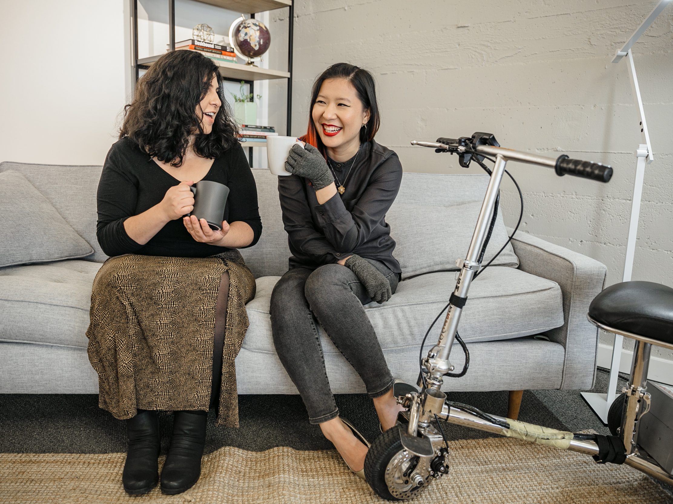 A Latinx disabled woman and an Asian disabled woman chat and sit on a couch, both holding coffee mugs. An electric lightweight mobility scooter rests on the side.