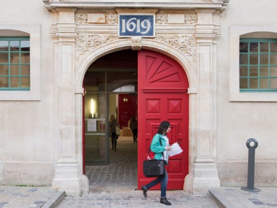 A female student exits a large red door with 1619 written above.