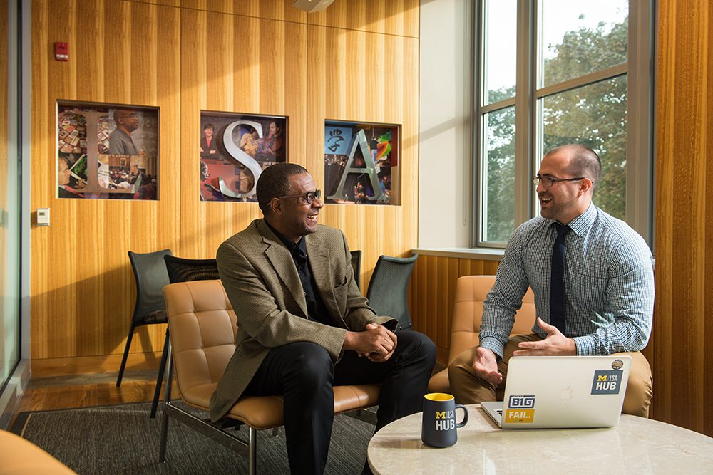 Ben Anderson engages in conversation with Harold Waters in an LSA meeting room; the letters "LSA" are on a wall decoration behind them and they are seated at a table with a Mac laptop on it.