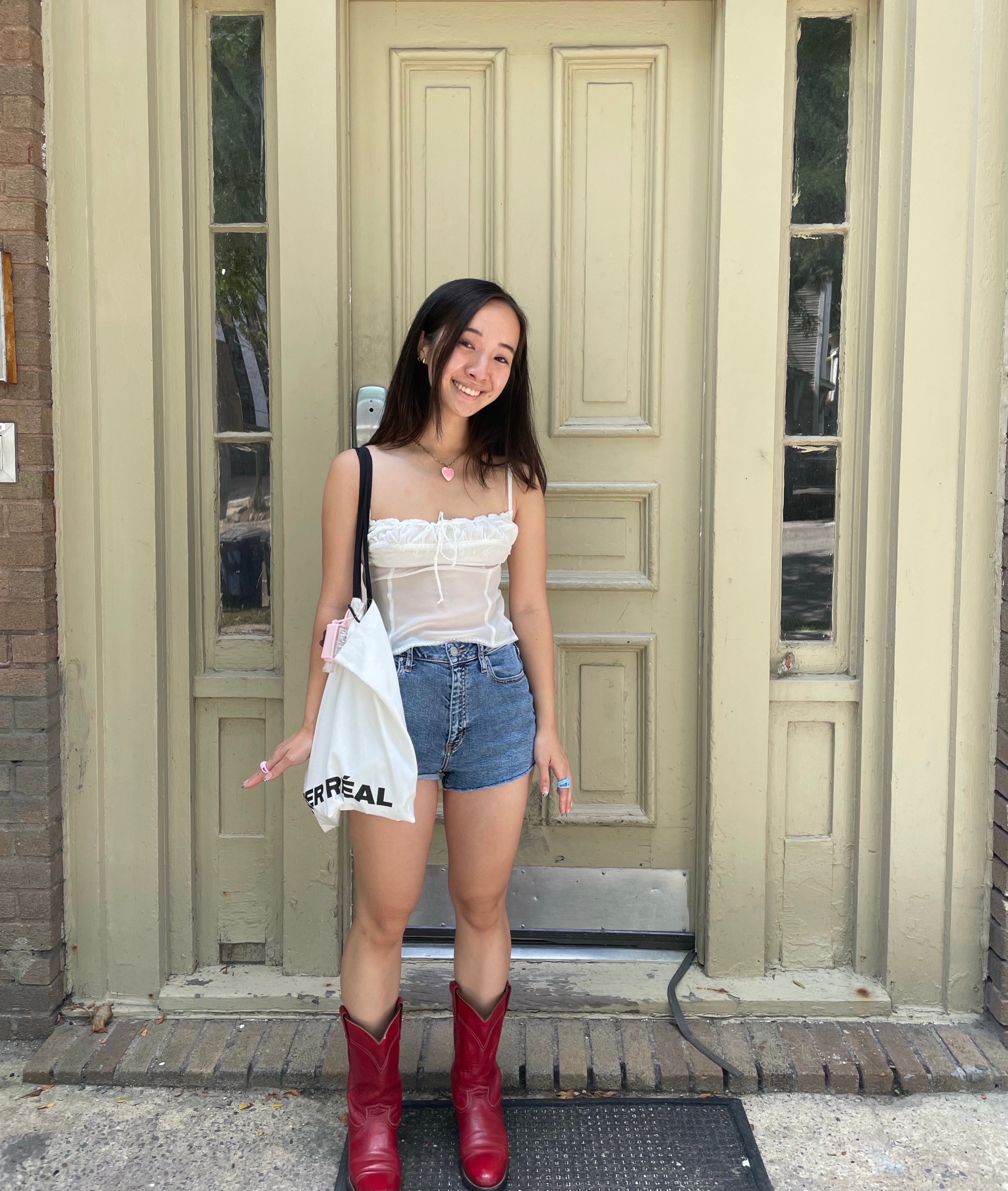Kathy smiling standing in front of a door, in a white top, jean shorts, and red cowboy boots