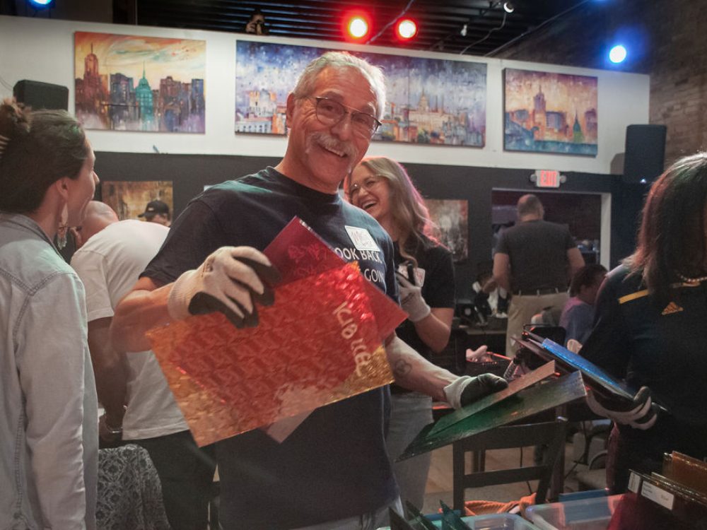Man in black T-shirt and mustache, holding sheets of colored glass with people behind him
