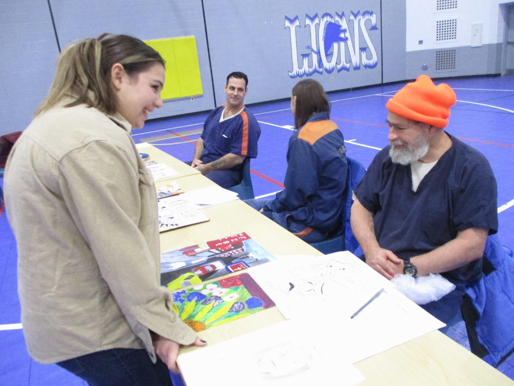 U-M student standing smiling and speaking to seated incarcerated artist with a table of paintings in between them.