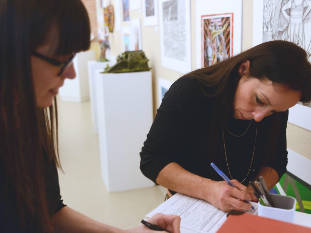 two exhibition guests signing a book at the gallery with art hanging on the walls behind them