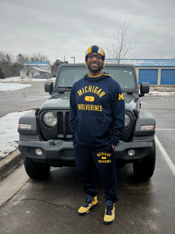 Man smiling in front of parked jeep wearing U-M sports apparel 