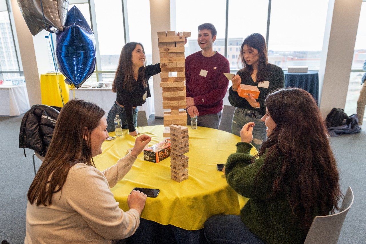 A group of students playing Jenga around a table with a maize tablecloth.