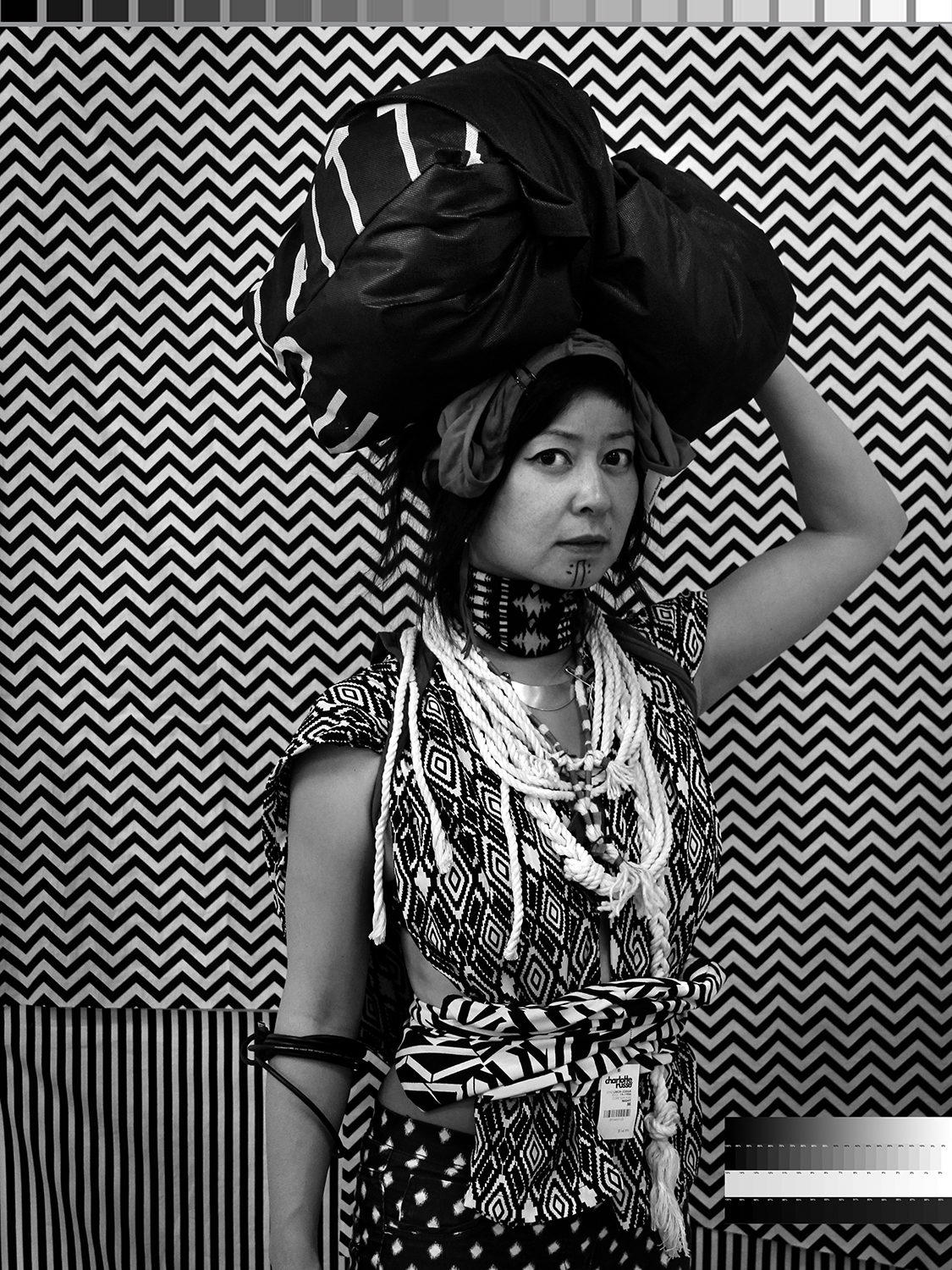 Woman in indigenous dress holding something on her head with a price tag on her clothes in black and white staring at the viewer