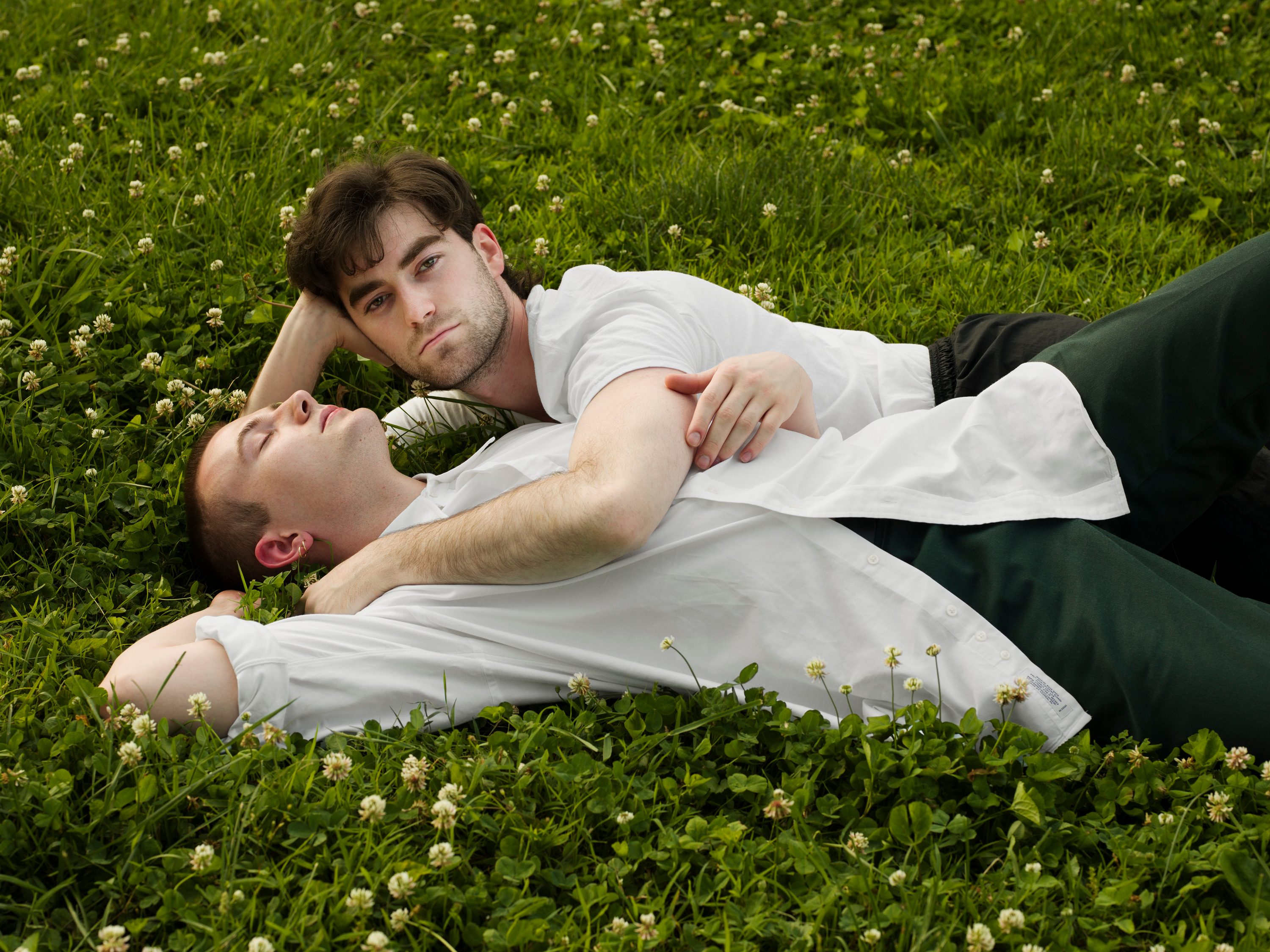 Two people relax in the grass. One person's head is back with closed eyes, the other looks at the camera.