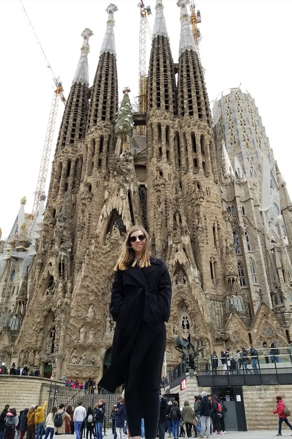 Katie Penrod stands in front of the Sagrada Familia, looking at the camera. Many people look up at the church in the background