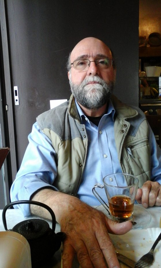 Javier Sanjines sits, holding a drink, wearing glasses and a beard, a blue collared shirt and a tan vest