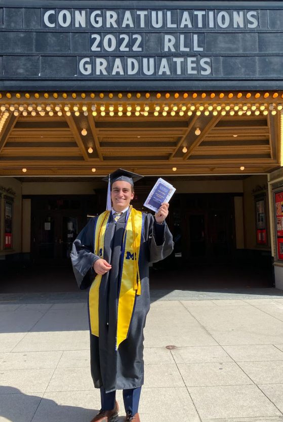 Image of UM Alum, Anthony DiBello (Italian Minor, 2022) standing in front of the Michigan Theater in Ann Arbor, MI. He is dressed in the traditional black cap and gown. The marquee above him says "Congratulations 2022 RLL Graduates".
