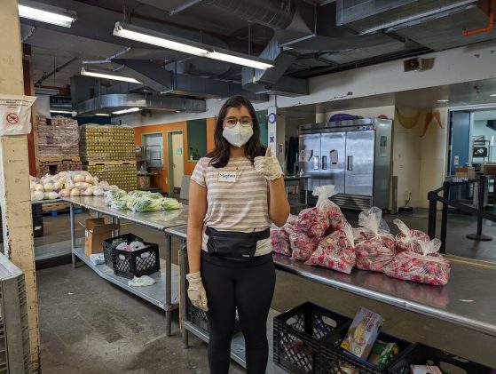 Image of Undergraduate Student, Meghna Singh (Spanish Major). She is wearing a face mask, striped shirt, and black pants; standing in an industry-grade kitchen prep area. She is packing food bags for distribution. 
