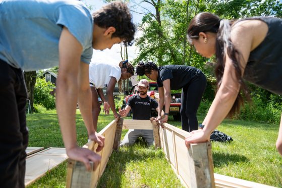 Five people help hold up three sides of a raised garden bed to connect them together. The grass is very green and they are surrounded by trees. 