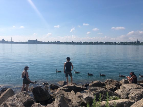 3 students stand on rocks with their backs turned at the edge of the still, blue Detroit River and a bright blue sky 