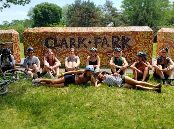 Students wearing helmets sit and lay on grass in front of a bright orange and red mosaic wall that reads "Clark Park"