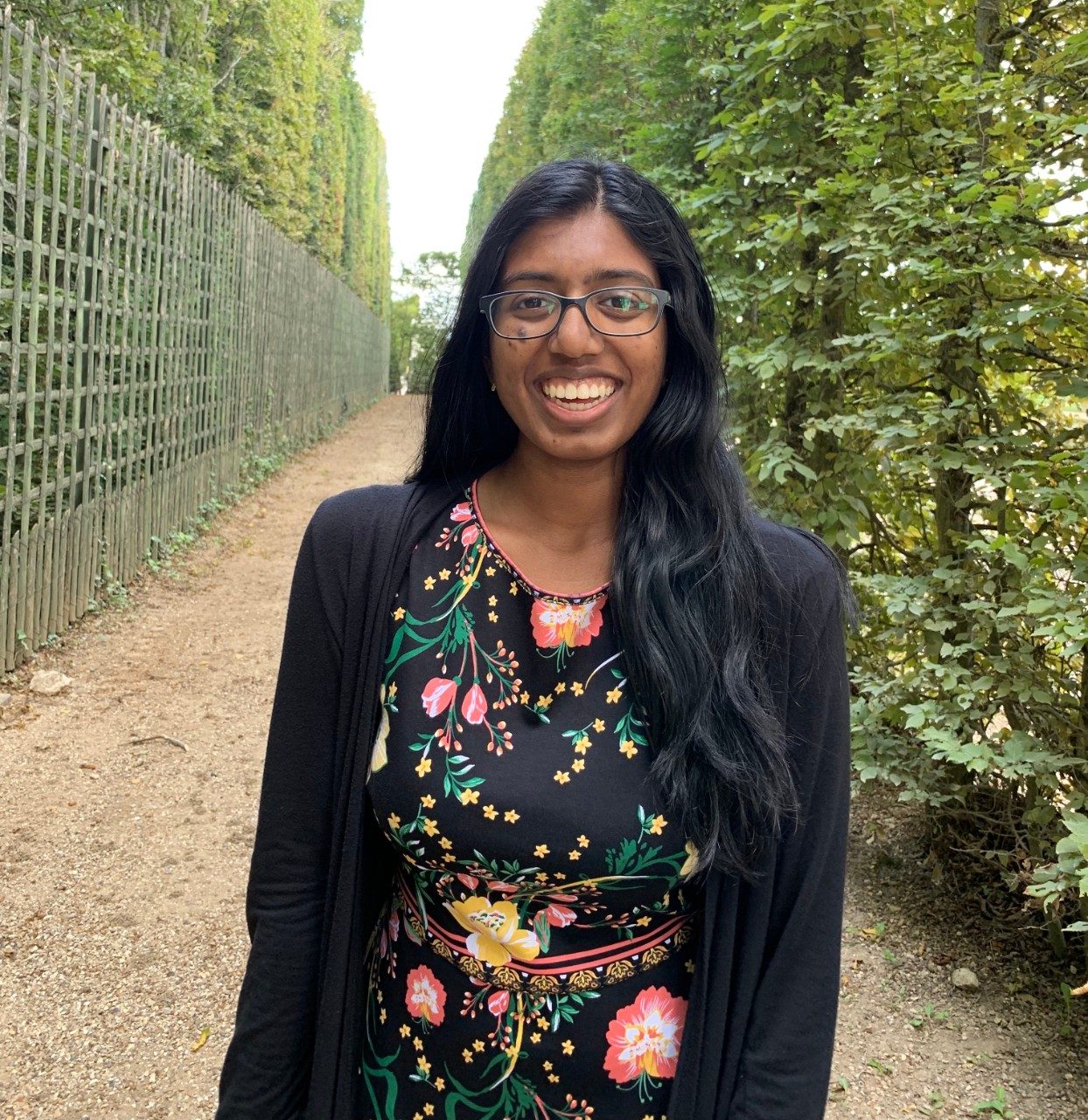 Meghna smiling center frame in front of a row of green trees