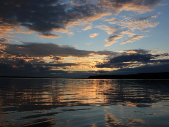 An early sunrise over an inland lake is shown, with a moody sky of blue clouds contrasting golden sun rays.