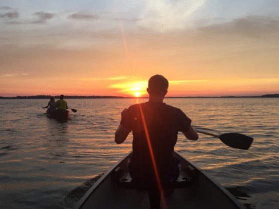 A man in a canoe paddles toward a beautiful sunset on a lake.