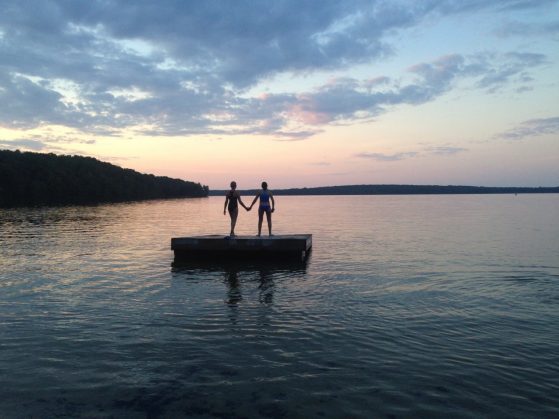 Two young women are show on a diving platform on an inland lake at sunset, with a sky of pinks and blues.
