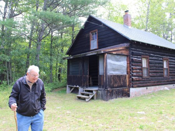 An elderly man with a walking stick stands outside a log cabin.