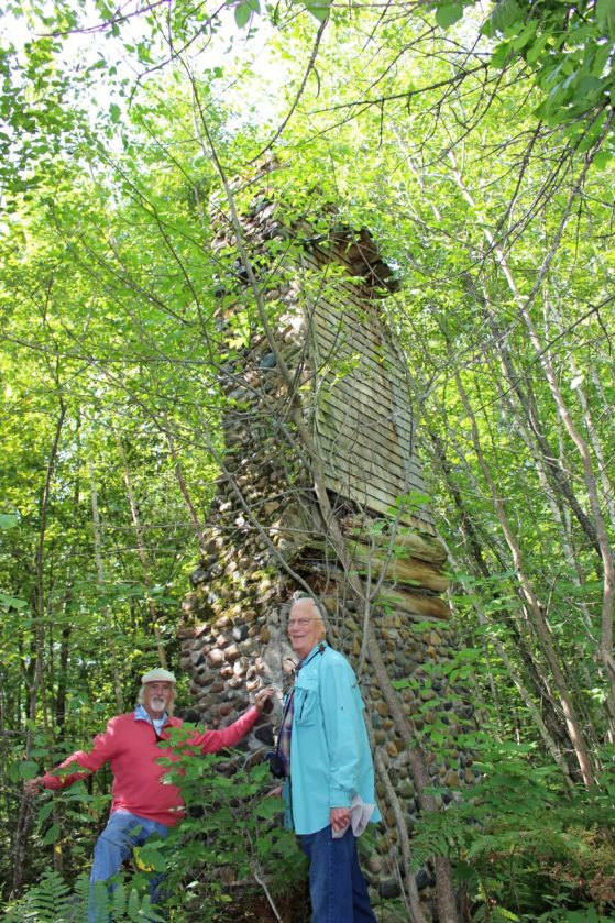 Two men stand next to a chimney, the ruins of a collapsed cabin in the forest. 