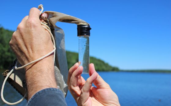 A pair of hands holds a small glass jar filled with lake water. In the background, a treed shoreline is visible.