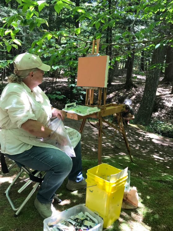 Cathy VanVoorhis sits before her easel and a fresh canvas, preparing to paint a wooded scene.