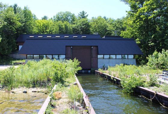 A building is shown from the water, with a brown metal roof and sliding garage door closing off a boat launch.