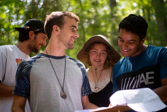 Smiling students flip through a lab packet in a verdant green forest. 