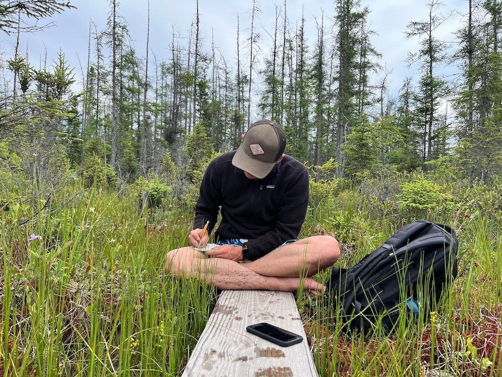 Student sitting on ground in forest, writing in a notepad on his lap