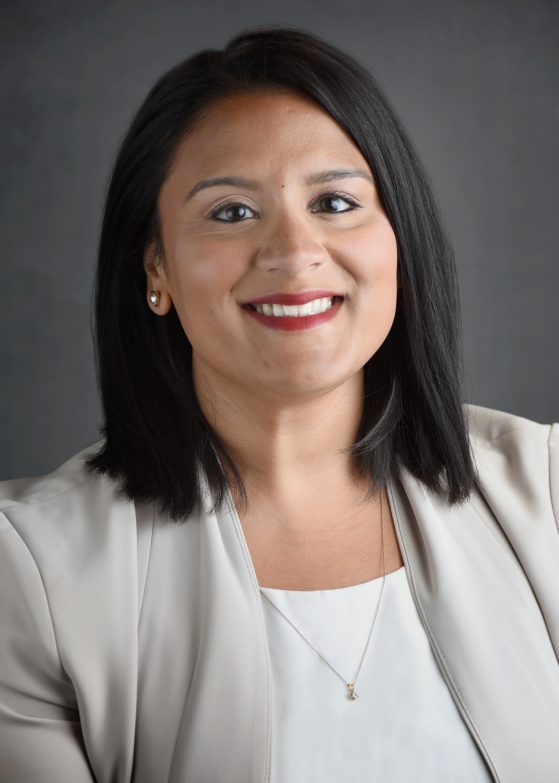 Nisha Pasupuleti has brown eyes, black straight hair to her shoulders, wearing a white shirt and tan blazer. sitting in front of a gray background.