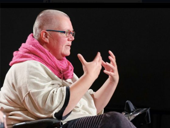 Bald headed person (Petra Kuppers) sits in wheelchair and gestures with hands during a performance on stage